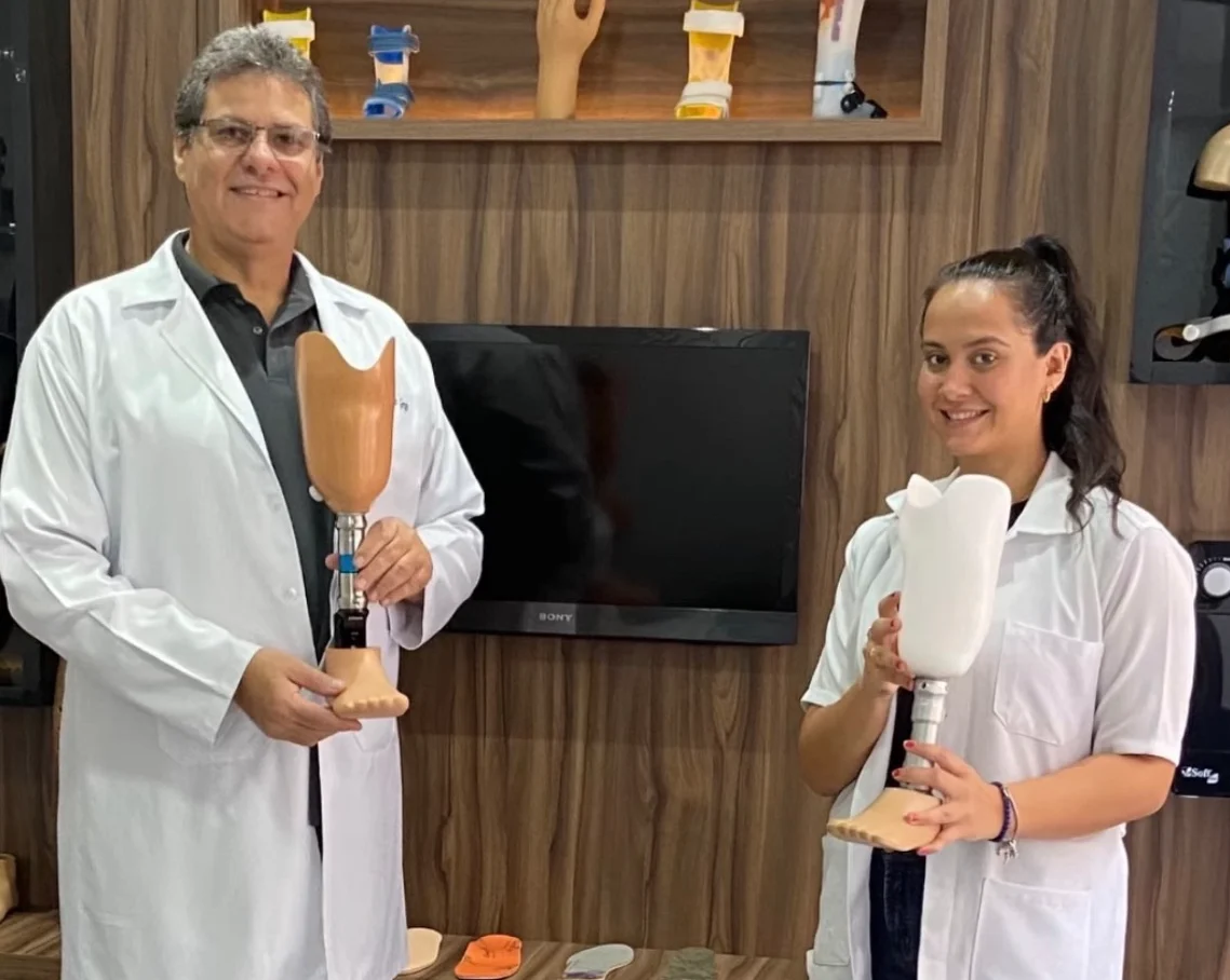 Dr. Henrique Grego, à esquerda, e Dra. Jéssica Maia, à direita, de jaleco branco na unidade da Futura Ortopedia, cada um segurando uma prótese de membro inferior